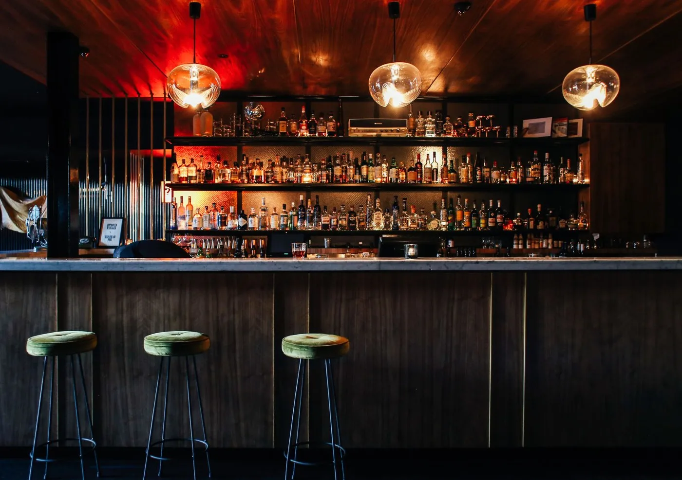 Barra de bar en madera oscura con taburetes y botellero iluminado